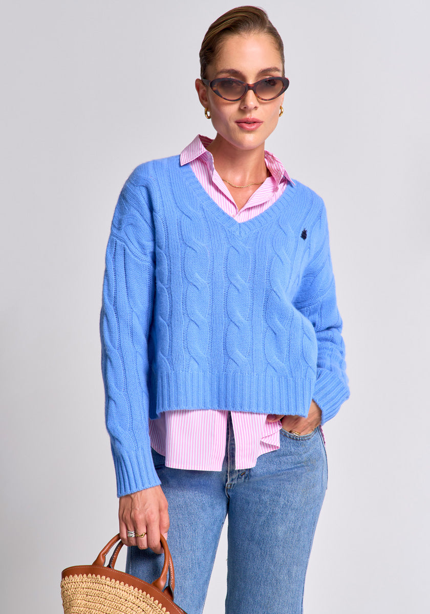 A woman in dark sunglasses and gold earrings poses in a blue Princess Goes Hollywood Zopfstrick Pullover Wolle Kaschmir over a pink striped shirt with jeans, holding a straw handbag, hair pulled back, against a plain light gray background.