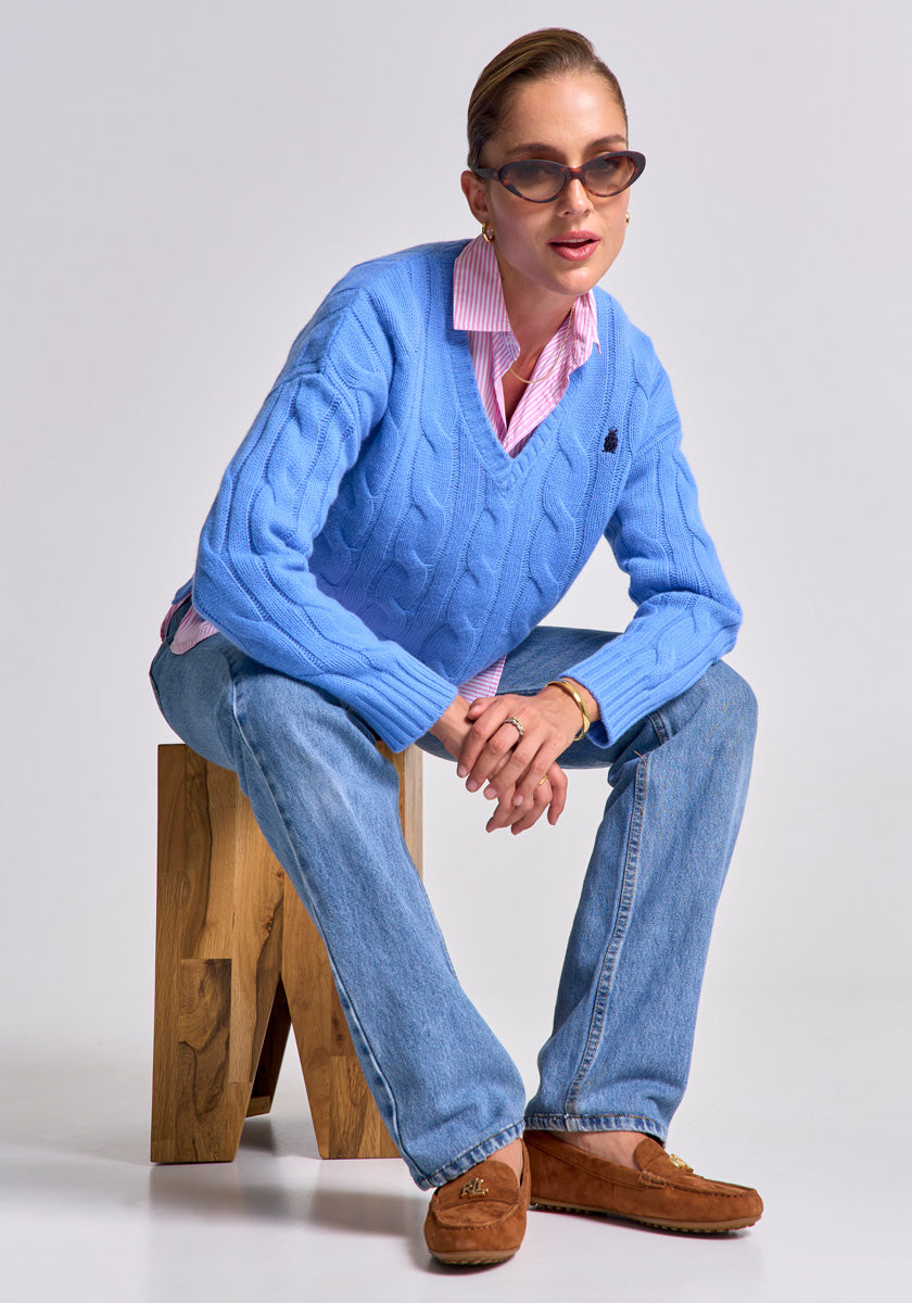 A woman in sunglasses, a Princess Goes Hollywood wool-cashmere pullover with cable knit over a pink collared shirt, blue jeans, and brown loafers sits on a wooden stool, leaning forward with arms on her knees against a plain studio background.