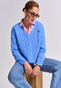A person with slicked-back hair and sunglasses wears a light blue v-neck cable-knit wool-cashmere sweater by Princess Goes Hollywood over a pink collared shirt with blue jeans, sitting on a wooden stool against a light gray background.