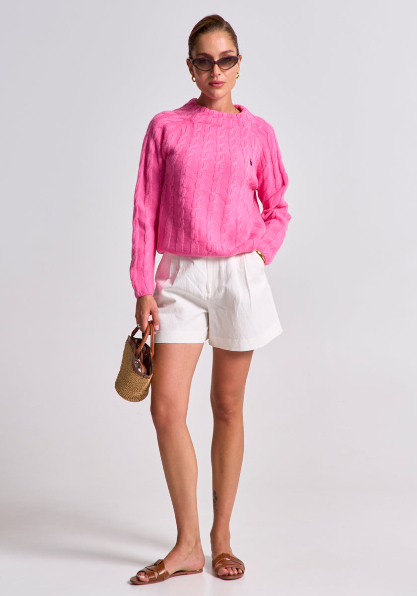 A woman stands against a plain background wearing sunglasses, a bright pink Princess Goes Hollywood wool-cashmere mix Zopfstrick pullover with Stehkragen, white high-waisted shorts, brown slide sandals, and holds a small woven handbag. Her hair is tied back.