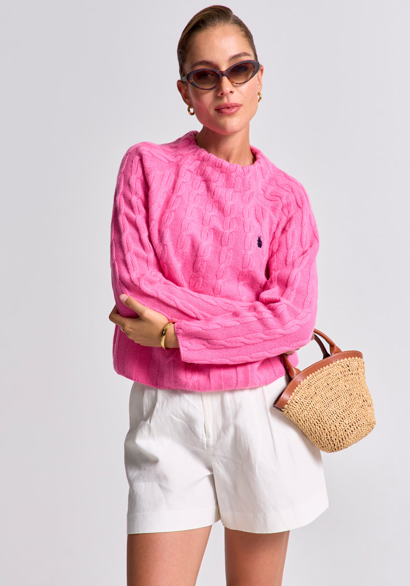A woman poses confidently in a bright pink Princess Goes Hollywood Zopfstrick Pullover and white shorts. With slicked-back hair, cat-eye sunglasses, gold earrings, and a woven straw handbag, she stands against a plain light gray background.
