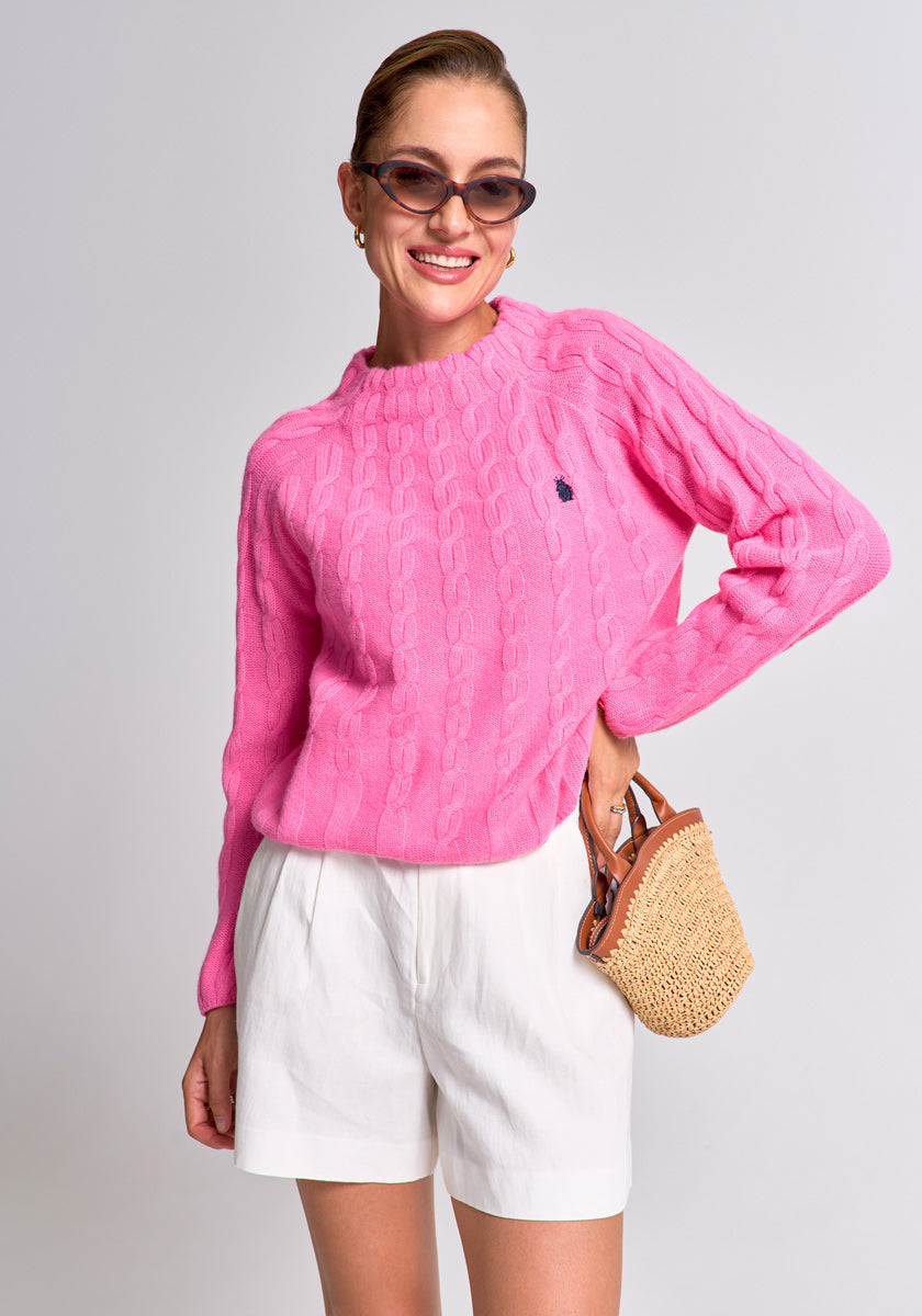 A smiling woman in cat-eye sunglasses wears a bright pink Princess Goes Hollywood Zopfstrick Pullover and white pleated shorts. She carries a small woven straw handbag, has slicked-back hair, and stands before a light gray background.