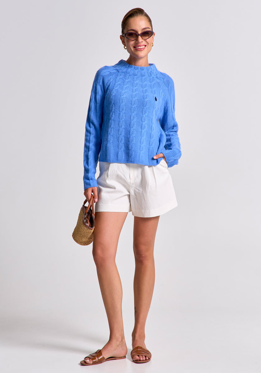 A woman smiles in a bright blue Princess Goes Hollywood Zopfstrick Pullover aus Wolle-Kaschmir-Mix, white shorts, and brown sandals. She holds a straw handbag, wears sunglasses, and poses with her light brown hair in a bun against a white background.