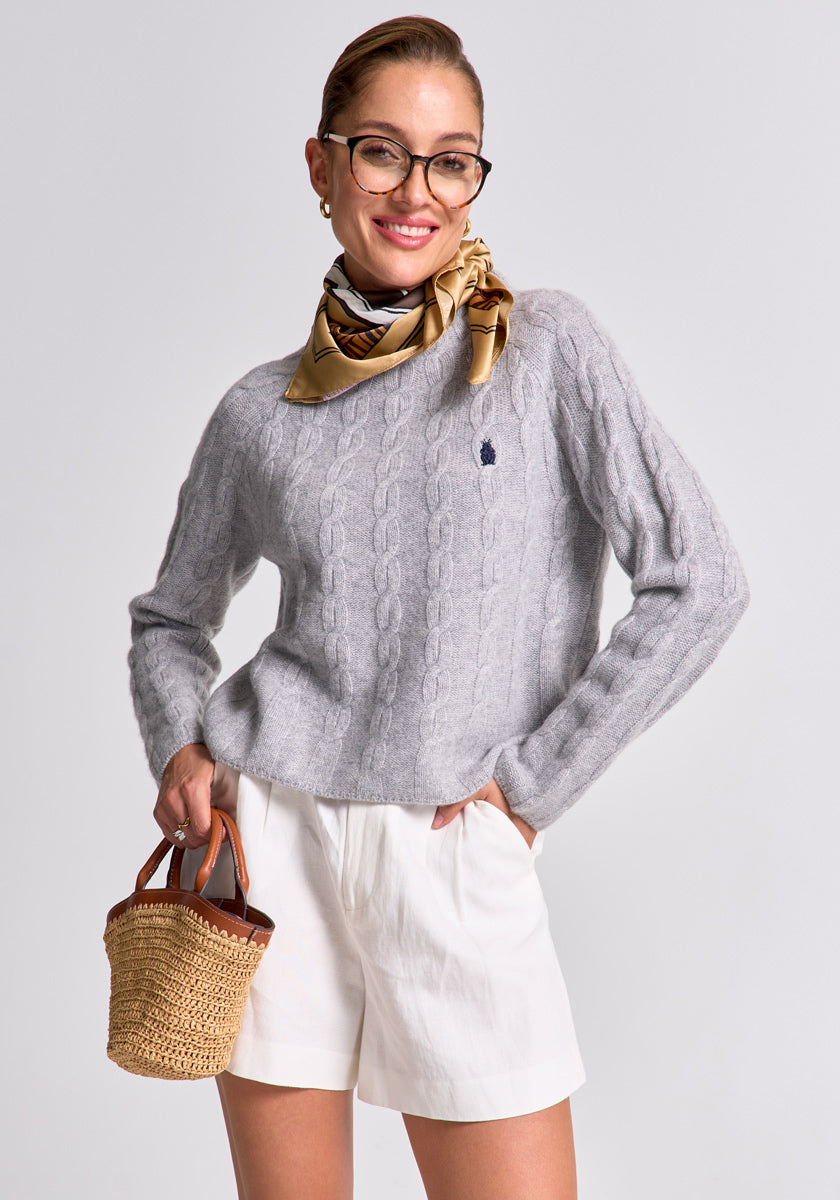 A smiling woman in glasses wears white shorts, a tan patterned scarf, and a light gray Princess Goes Hollywood Zopfstrick Pullover aus Wolle-Kaschmir-Mix mit Stehkragen. She stands against a plain background, holding a woven straw handbag with one hand in her pocket.