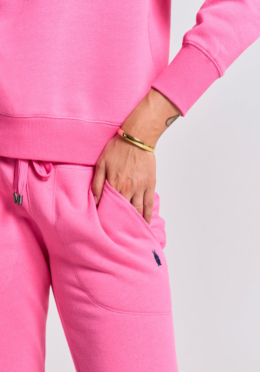 Wearing a bright pink Princess Goes Hollywood Baumwolle Sweatpullover and matching drawstring sweatpants with a blue logo, the person puts their right hand in the pocket. A thin gold bracelet and a small forearm tattoo are visible.