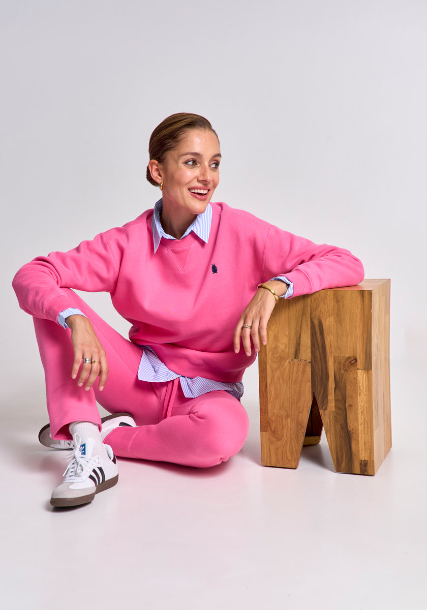 A woman with pulled-back hair sits on the floor, leaning on a wooden block. She wears bright pink Princess Goes Hollywood Regular Fit Baumwoll Jogginghose with fleece lining over a blue collared shirt and white Adidas sneakers, smiling to the side.