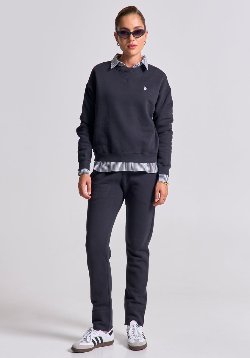 A woman stands against a plain background, wearing sunglasses, her hair pulled back. She wears a black Princess Goes Hollywood Baumwolle Sweatpullover over a collared shirt with striped cuffs, matching black pants, and white Adidas sneakers with black stripes.