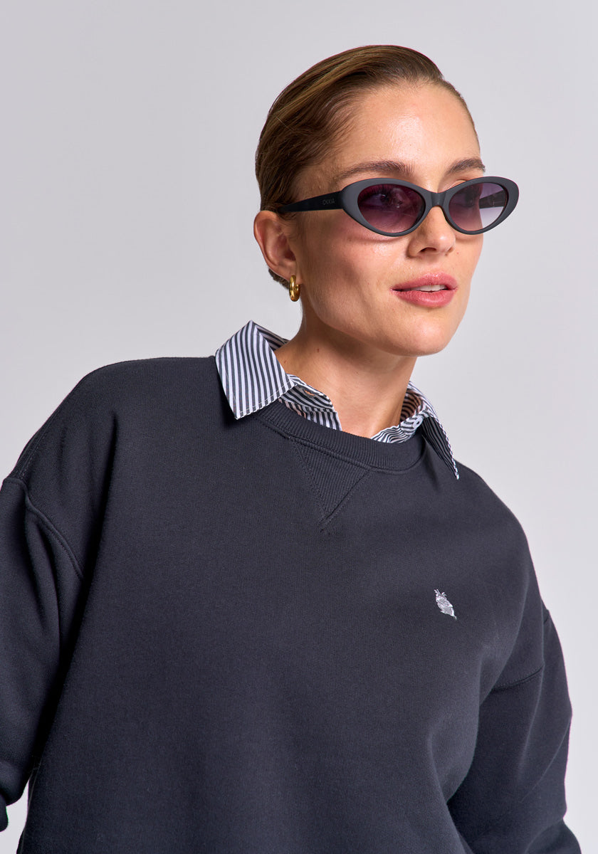 A woman with short, slicked-back hair wears Princess Goes Hollywood black cat-eye sunglasses, a gold hoop earring, and a black Baumwolle Sweatpullover with an embroidered logo over a blue-and-white striped shirt against a light gray background.