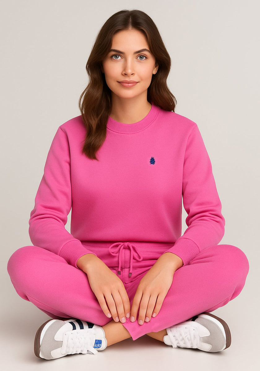 A woman with long brown hair sits cross-legged on the floor, smiling softly and facing forward. She wears a bright pink Princess Goes Hollywood Regular Fit Jogginghose and sweatshirt set made of soft Baumwolle against a plain light background.