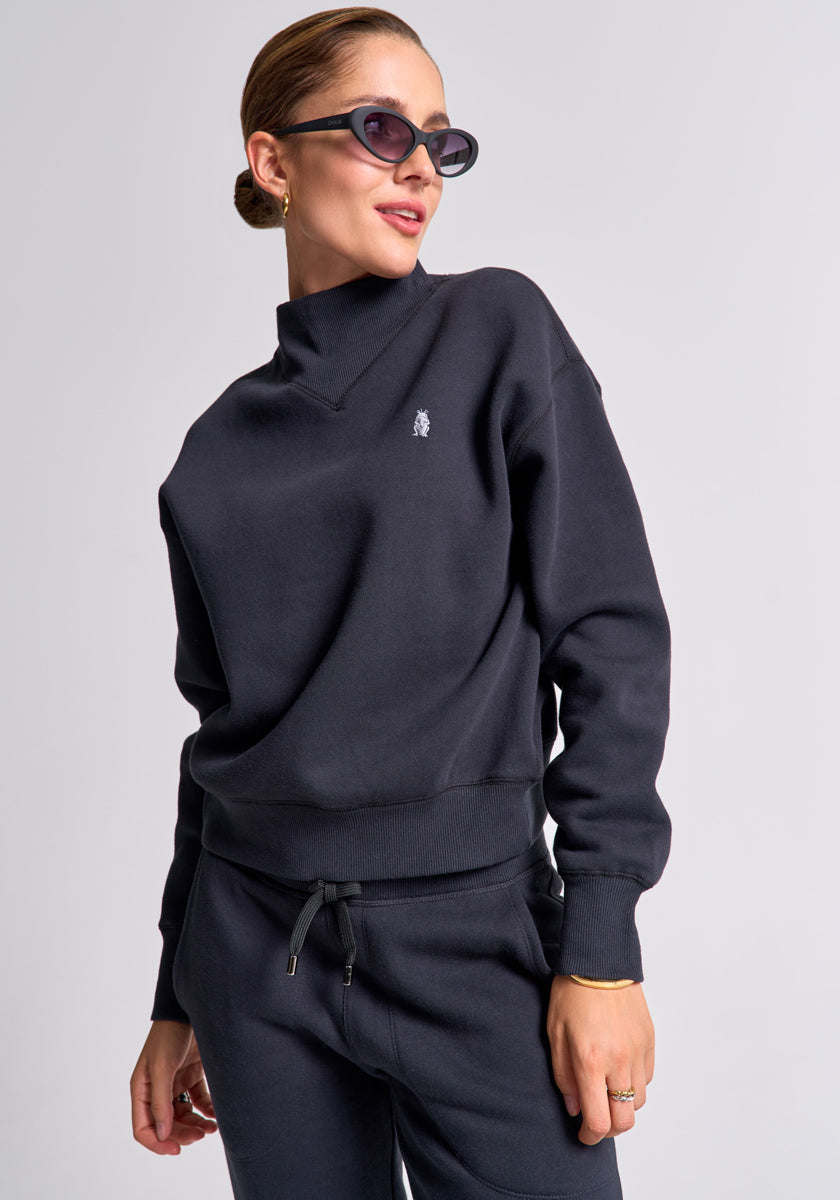 A woman in sunglasses and gold jewelry models a black fleece Sweatpullover and matching joggers from Princess Goes Hollywood, featuring a small embroidered chest logo. She poses against a plain light gray background.