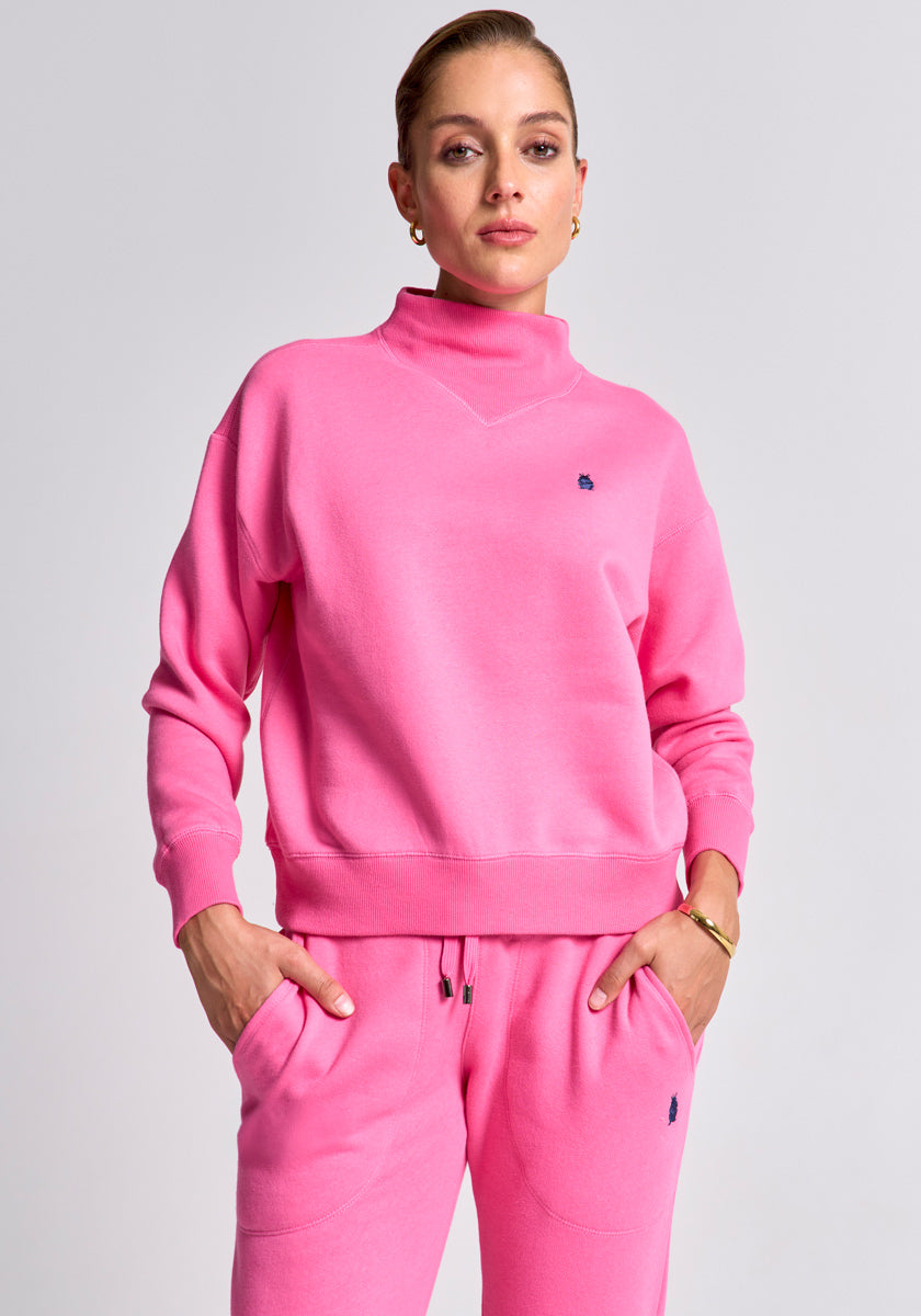 A woman faces the camera wearing a bright pink Princess Goes Hollywood Baumwolle Sweatpullover and matching sweatpants. She has short, slicked-back hair, gold hoop earrings, and a bracelet, with her hands in her pockets against a light gray background.