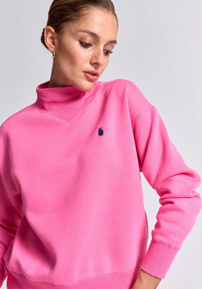 A light-skinned woman with slicked-back hair wears a bright pink Princess Goes Hollywood Baumwolle Sweatpullover with a small dark blue embroidered chest logo, gazing downward to her left against a plain light gray background.