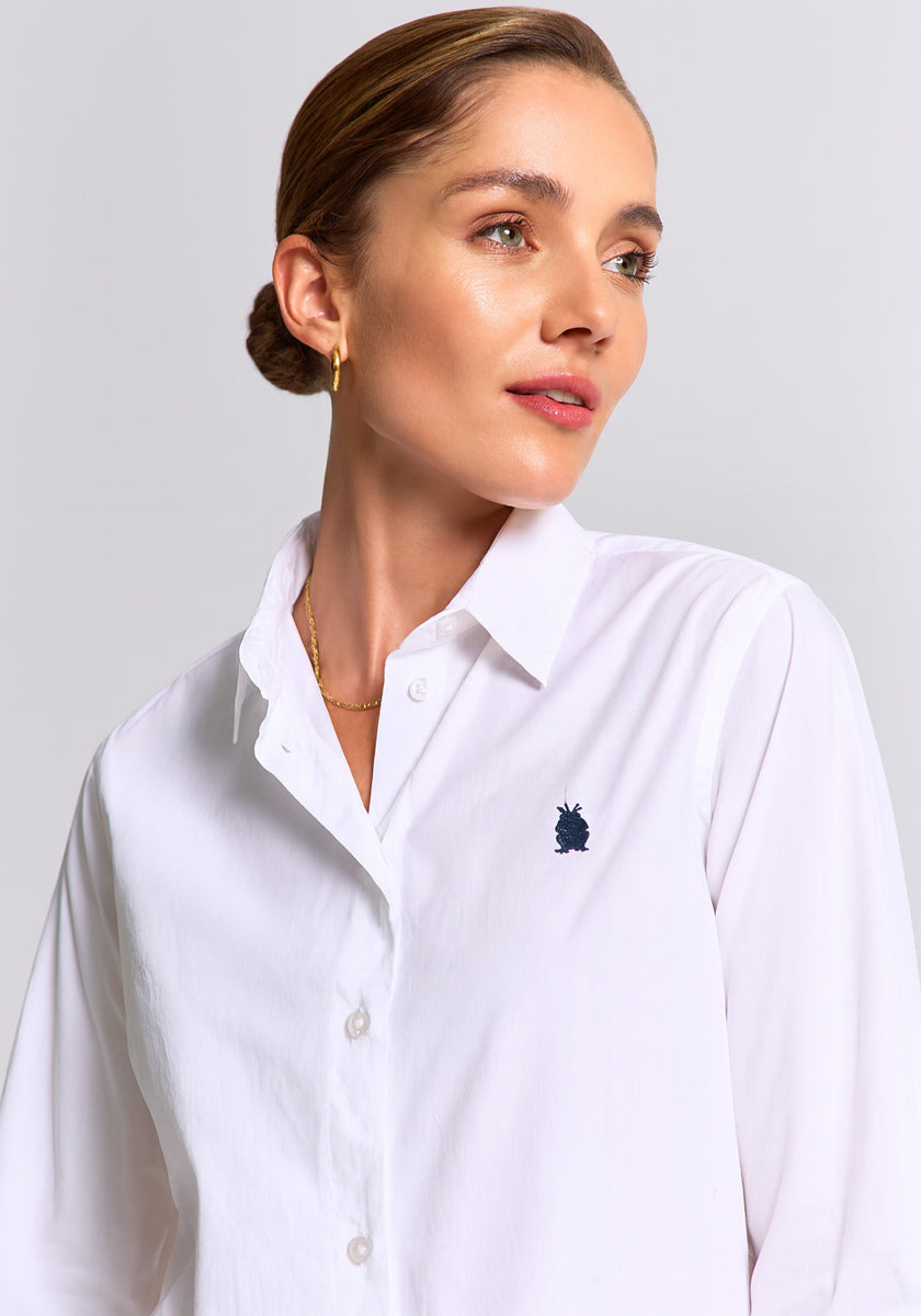 A woman with light skin and swept-back brown hair wears a Princess Goes Hollywood white Baumwollhemd in Regular Fit, featuring a small dark blue chest logo. She pairs it with gold hoop earrings and a necklace, gazing slightly left against a plain background.