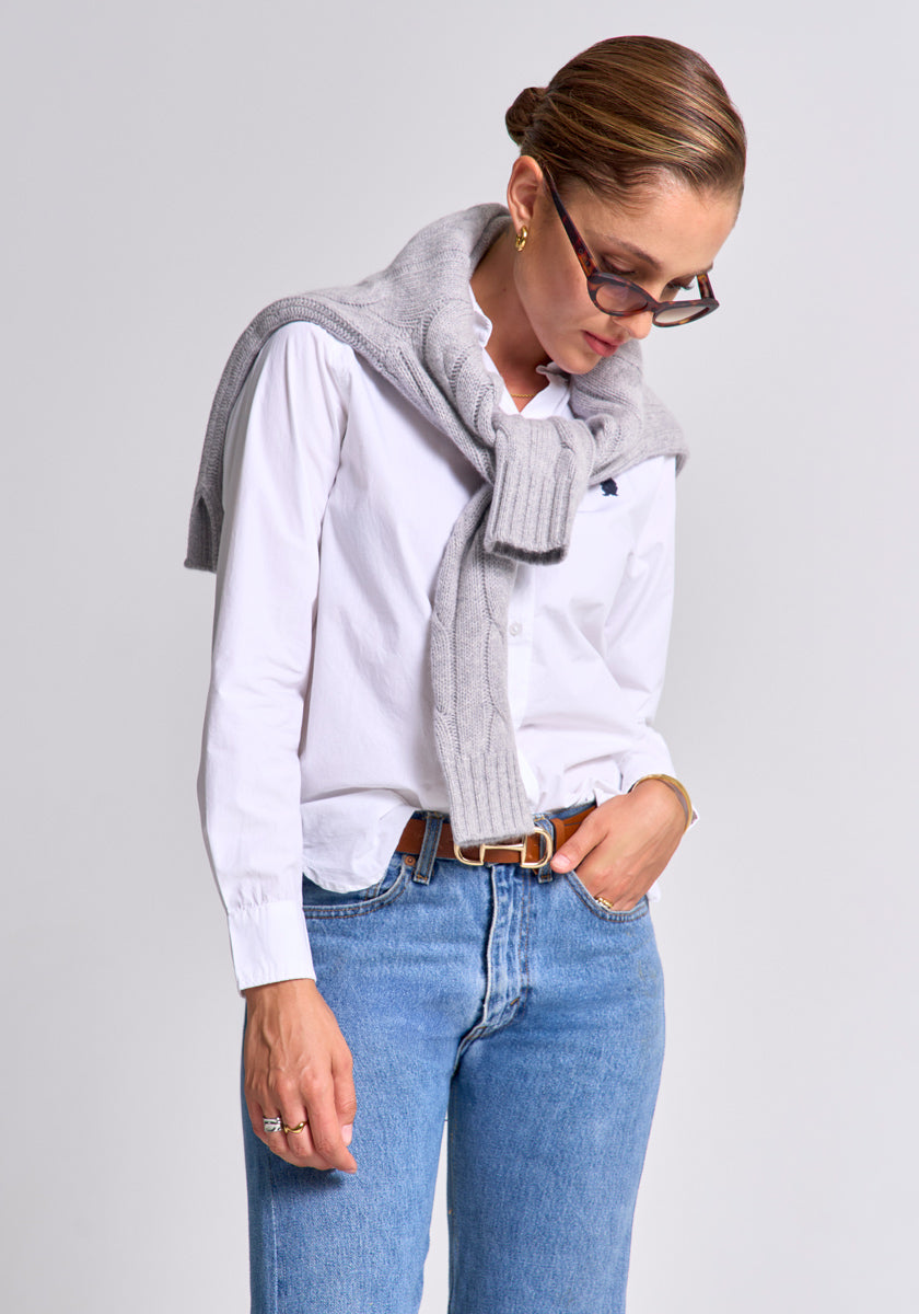 A woman with brown hair in a bun, wearing glasses, a white button-up shirt, blue jeans, and a light gray Princess Goes Hollywood wool-cashmere V-neck sweater draped over her shoulders, looks down to the side with one hand in her pocket.