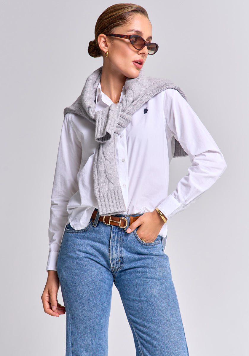 A woman exudes confidence in a Princess Goes Hollywood grey wool-cashmere sweater draped over her shoulders, styled with gold earrings, blue jeans, a white button-down shirt, sunglasses, and slicked-back hair against a plain background.