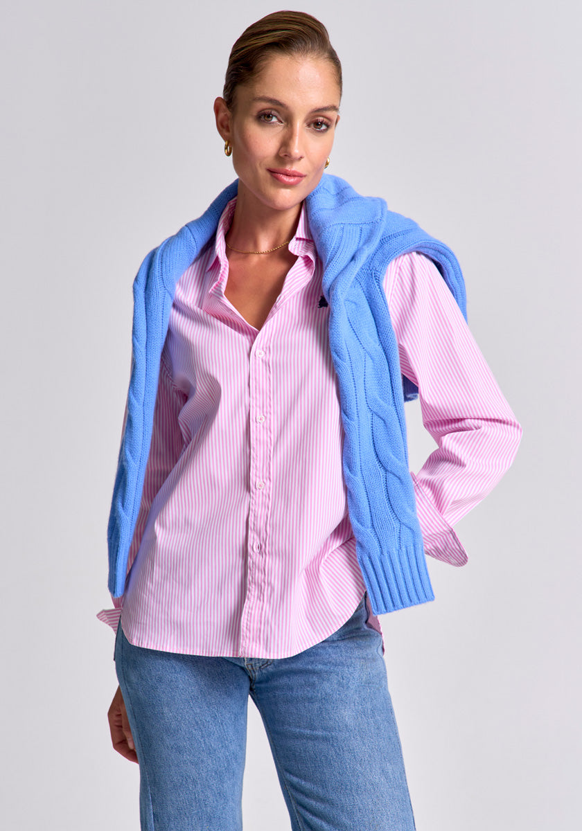A woman stands confidently against a light gray background, wearing a light pink striped button-up shirt, blue jeans, and a Princess Goes Hollywood Zopfstrick Pullover Wolle-Kaschmir mit V-Ausschnitt locker über die Schultern gelegt. Her hair is pulled back.