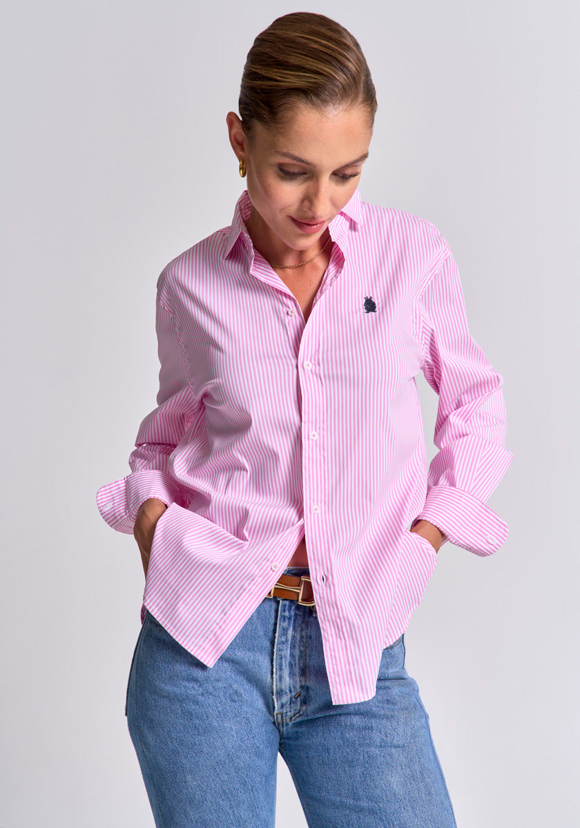 A woman with light brown hair pulled back wears a pink and white striped Princess Goes Hollywood Regular Fit cotton shirt with a navy logo on the chest. She has her hands in blue jeans pockets and looks down against a plain light background.