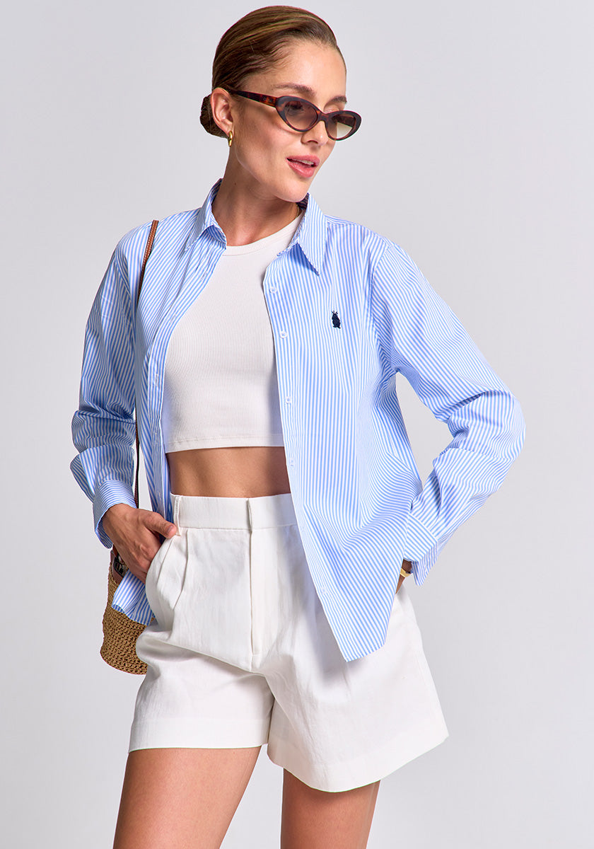 A woman poses confidently in Princess Goes Hollywood cat-eye sunglasses, a blue-striped Regular Fit Baumwollhemd with Polokragen over a white crop top, high-waisted white shorts, and carries a straw bag. Her hair is tied back against a plain light gray background.