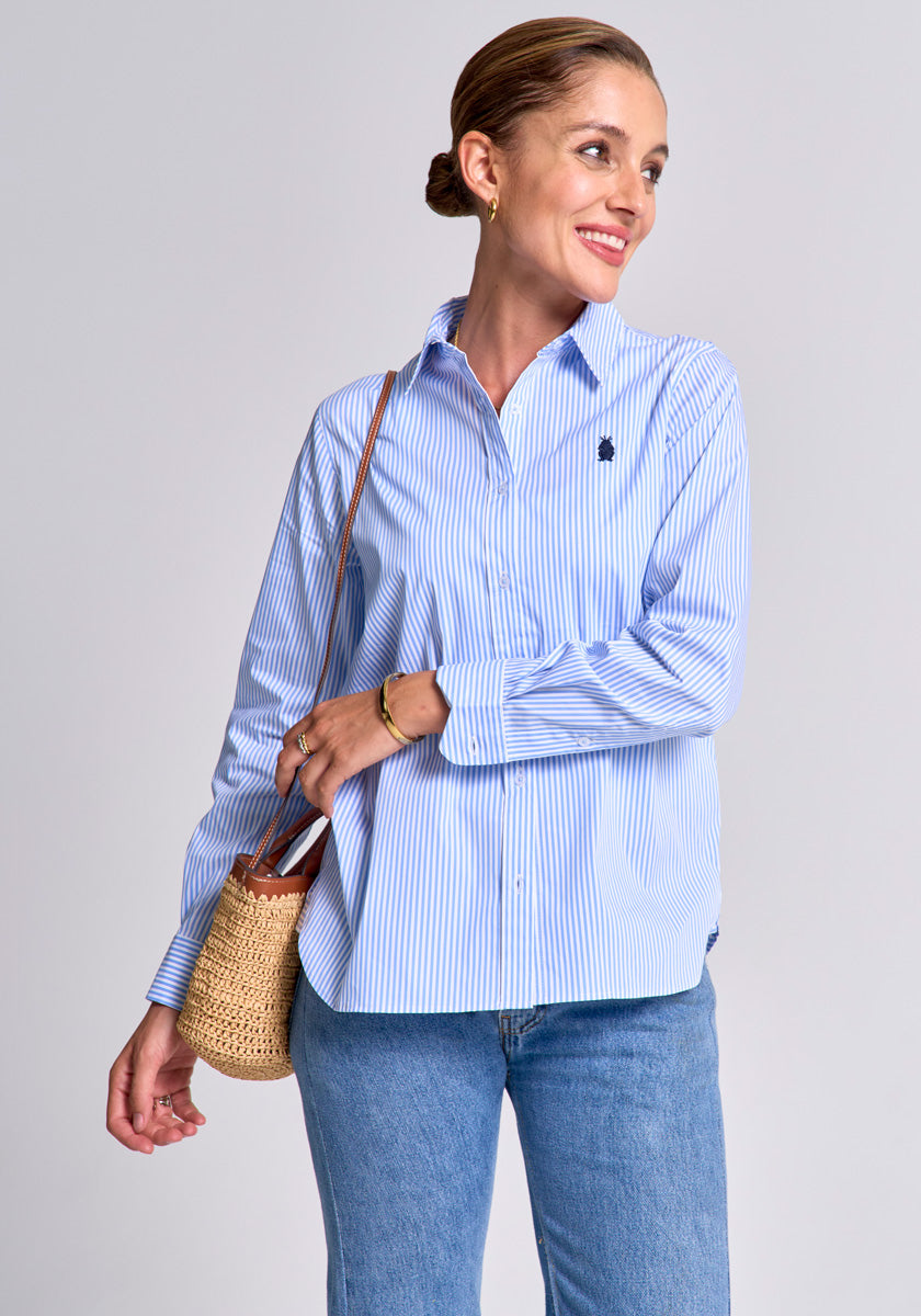 A woman stands against a gray background, smiling and looking to the side. She wears a regular fit gestreiftes Baumwollhemd with Polokragen from Princess Goes Hollywood, blue jeans, hoop earrings, and carries a woven straw handbag.