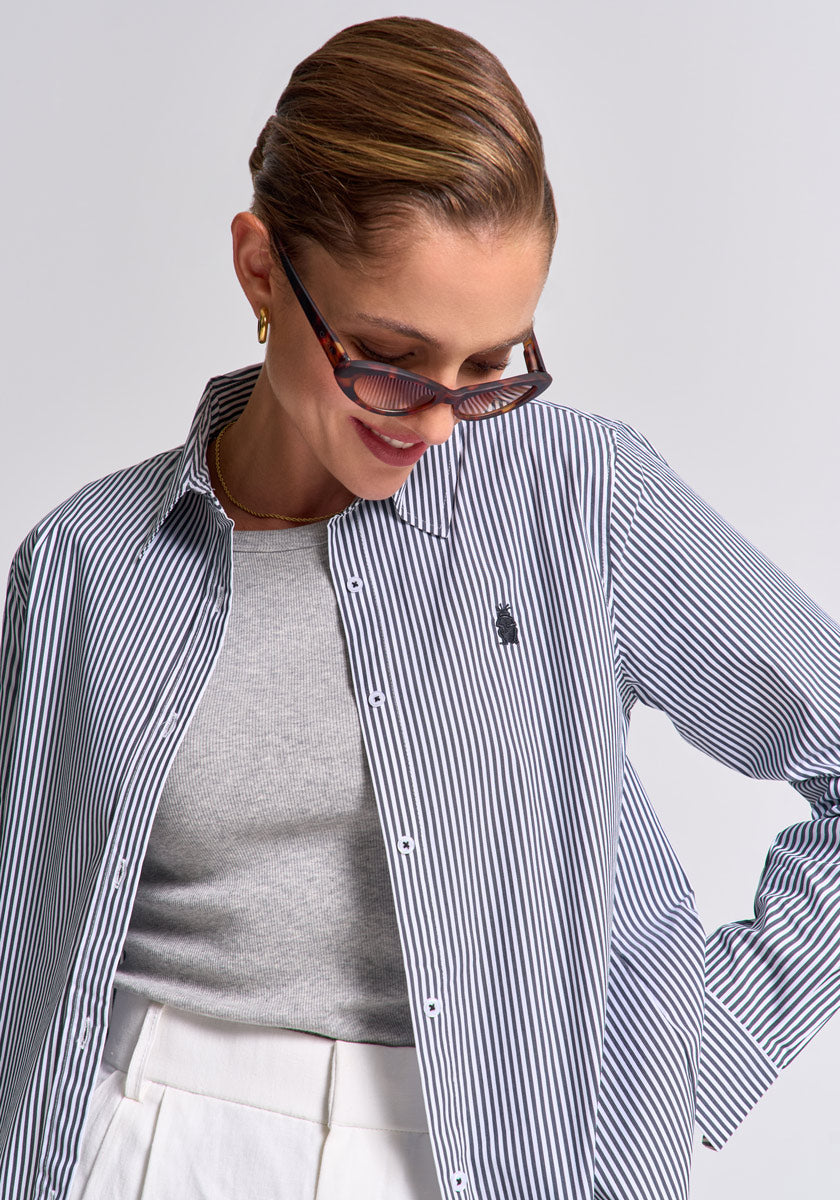 A woman with slicked-back hair wears Princess Goes Hollywood tortoiseshell sunglasses, a gray top, a striped cotton polo shirt, and high-waisted white pants. She looks down and smiles softly against a neutral background.
