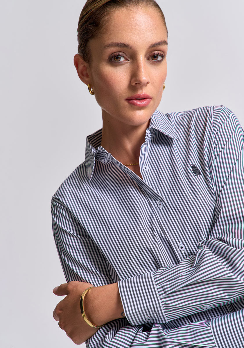 A woman with slicked-back hair wears a gestreiftes Baumwollhemd in Regular Fit mit Polokragen von Princess Goes Hollywood, goldenen Creolen und einem dünnen Armreif. Sie blickt selbstbewusst vor grauem Hintergrund, die Arme locker verschränkt.