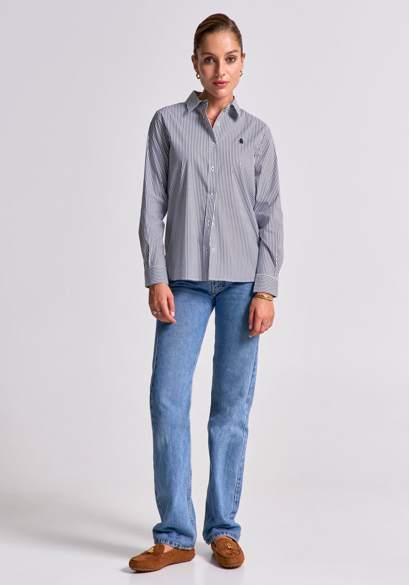 A woman stands against a plain light background in a blue-and-white striped cotton shirt with polo collar by Princess Goes Hollywood, light blue jeans, and brown moccasin-style shoes. Her hair is pulled back and her expression is neutral.