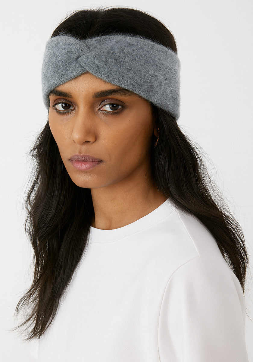 A woman with long, dark hair wears a Princess Goes Hollywood hellblau-graues Kaschmir-Stirnband mit Twistdetail. She has a neutral expression and is dressed in a plain white top against a plain white background.
