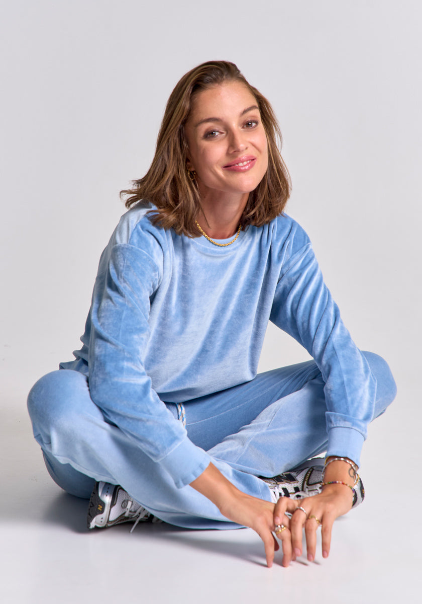A woman with shoulder-length brown hair sits cross-legged, smiling at the camera in a light blue Princess Goes Hollywood tracksuit and white sneakers, accessorized with bracelets and a necklace against a plain, light background.