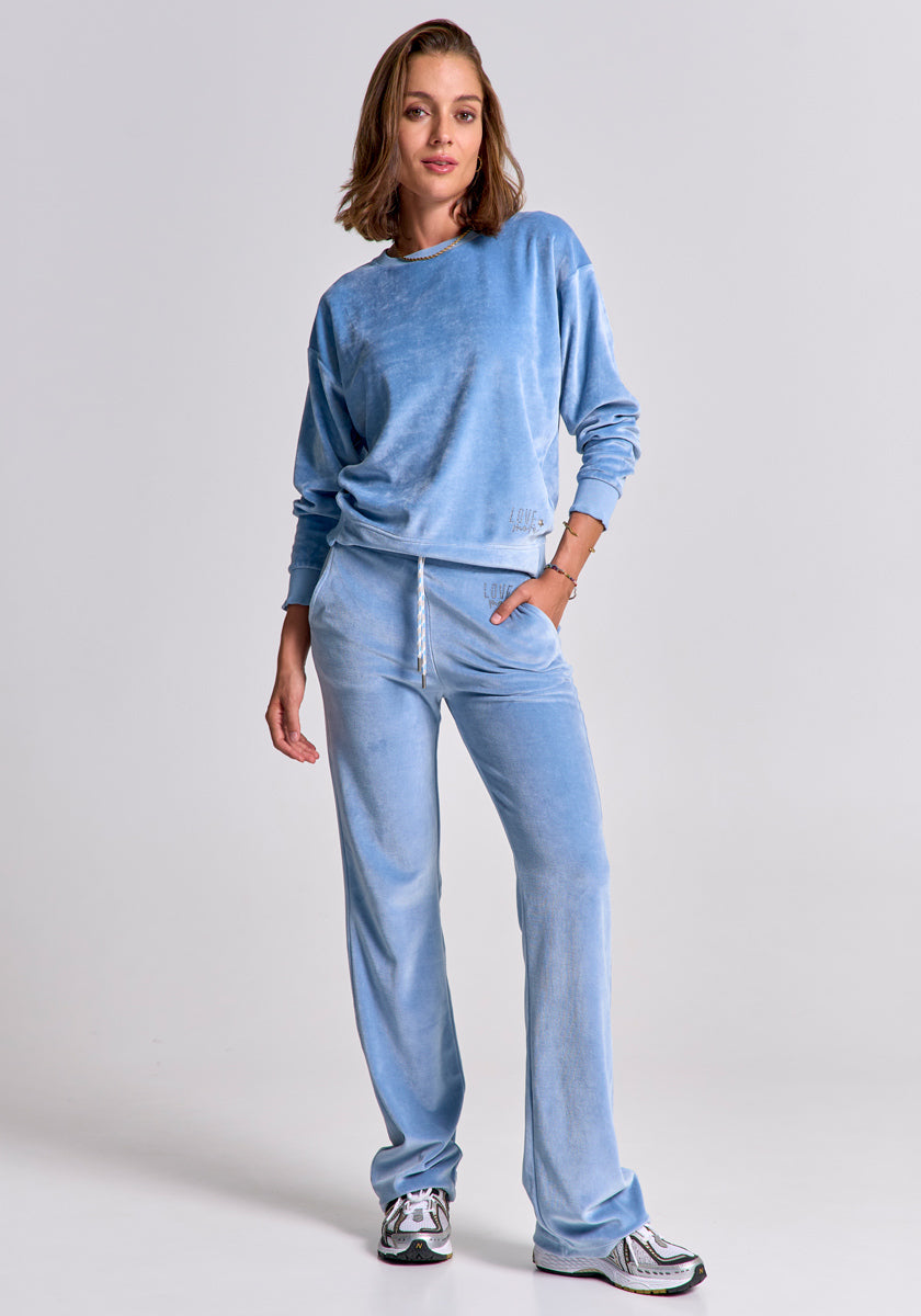 A woman with short brown hair stands against a plain background in a light blue Princess Goes Hollywood tracksuit with long sleeves and drawstring sweatpants. She puts one hand in her pocket, wearing white and silver sneakers, exuding confidence.