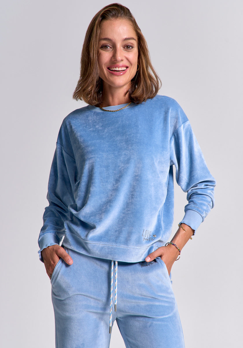 A woman with shoulder-length light brown hair smiles at the camera, wearing a light blue Princess Goes Hollywood Sweatshose aus Nicki-Stoff and matching sweatshirt. One hand in her pocket, she stands against a plain background that highlights her comfy style.