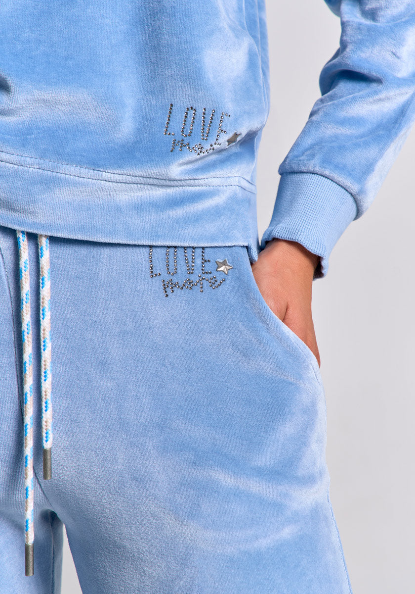 A person wears a light blue Princess Goes Hollywood Nicki-Stoff tracksuit with white and blue drawstrings. Both Sweatshose and sweatshirt show rhinestone "LOVE more" text with a small star gem. Their right hand is in their pocket; background is plain gray.