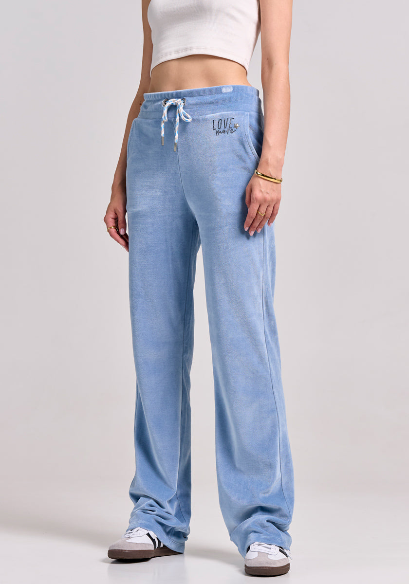 Someone stands against a plain background wearing light blue Princess Goes Hollywood Sweatshose aus Nicki-Stoff with a drawstring waist and “LOVE” graphic on the hip, paired with a white crop top, white sneakers, and a gold bracelet. Their face is not visible.