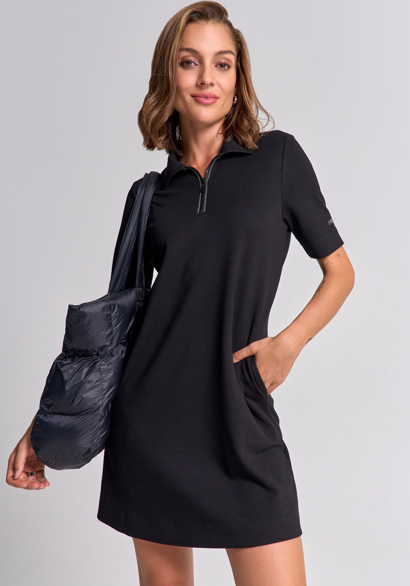 A woman with chin-length brown hair wears a black Mini Sweatkleid by Princess Goes Hollywood featuring a partial front zipper and short sleeves. She carries a puffy black tote, poses with one hand in her pocket, and smiles slightly against a plain background.