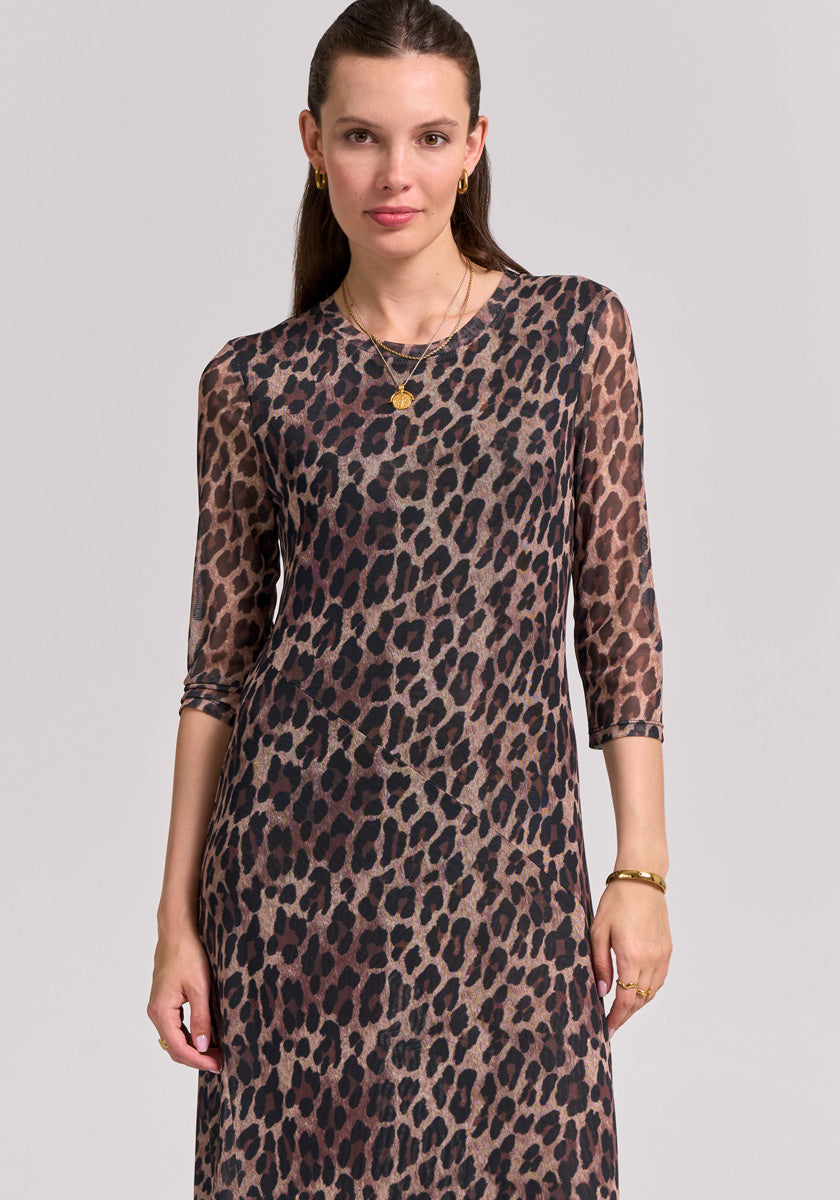 A woman with light skin and long brown hair pulled back wears a fitted Mesh Kleid by Princess Goes Hollywood featuring a bold Glam-Leo-Print, accessorized with gold hoop earrings, layered necklaces, and a bracelet against a light gray background.