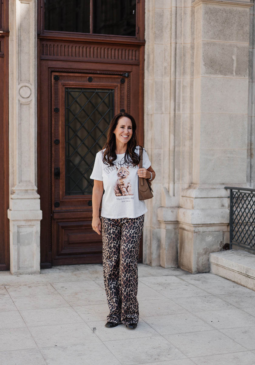 A woman with long dark hair smiles in front of a wooden door, wearing a Princess Goes Hollywood weißes Baumwoll T-Shirt mit Rundhalsausschnitt, loose Luchs-Print pants, black shoes, and carrying a brown shoulder bag.