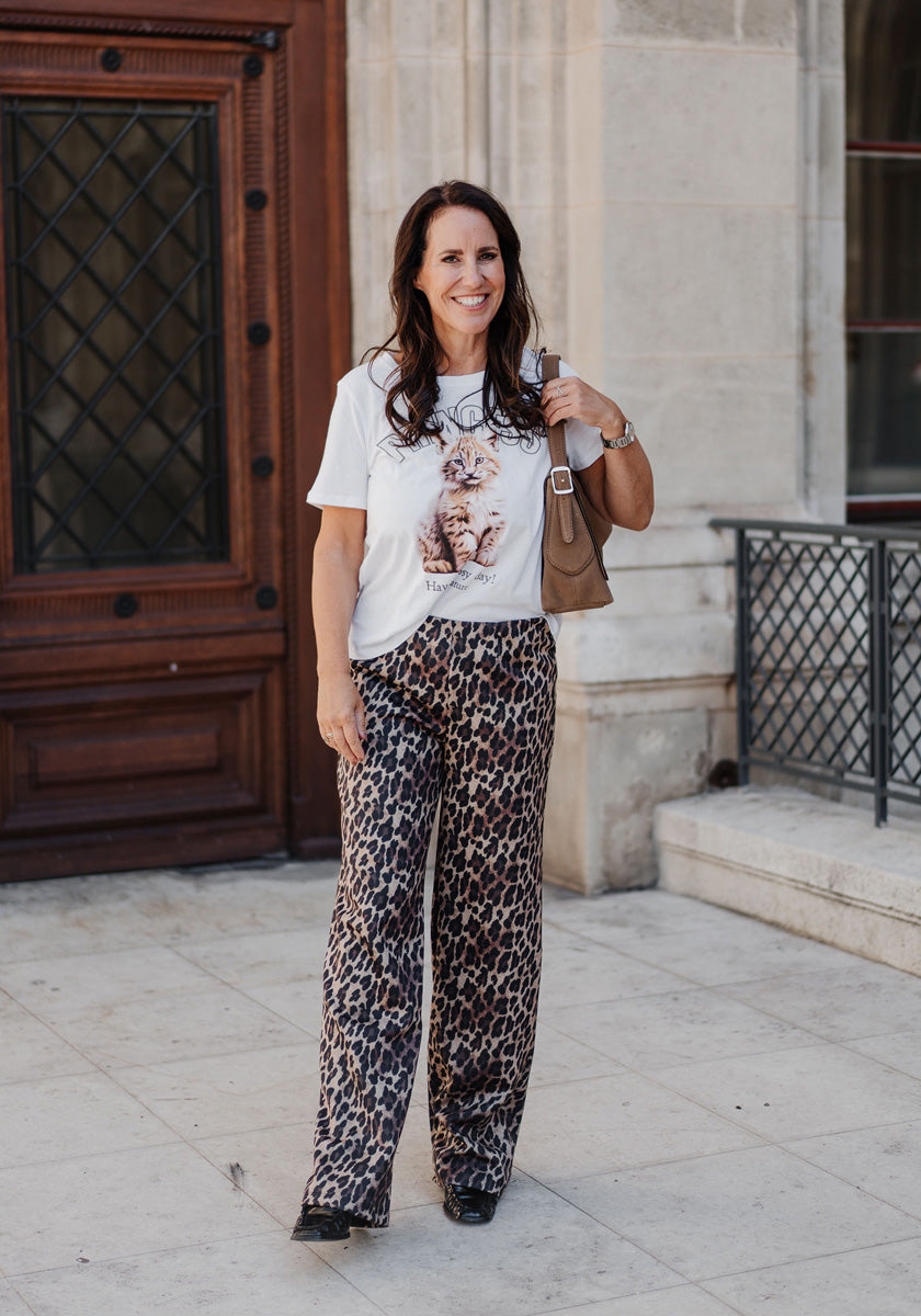 A woman with long dark hair smiles outdoors, wearing a Princess Goes Hollywood white Baumwoll T-Shirt mit Rundhalsausschnitt with a lion graphic, loose Luchs-Print pants, black shoes, and carrying a brown handbag by a stone building.