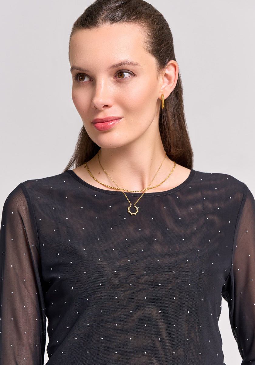 A woman with straight brown hair pulled back wears gold hoop earrings and layered necklaces—one with a geometric pendant—over a Princess Goes Hollywood black mesh shirt with sheer sleeves and delicate rhinestone accents. She smiles softly, facing right.
