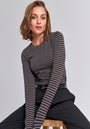 A woman with chin-length brown hair sits confidently on a wooden stool, wearing a Princess Goes Hollywood striped long-sleeve stretch shirt tucked into high-waisted black pants and a black belt, set against a plain light gray background.