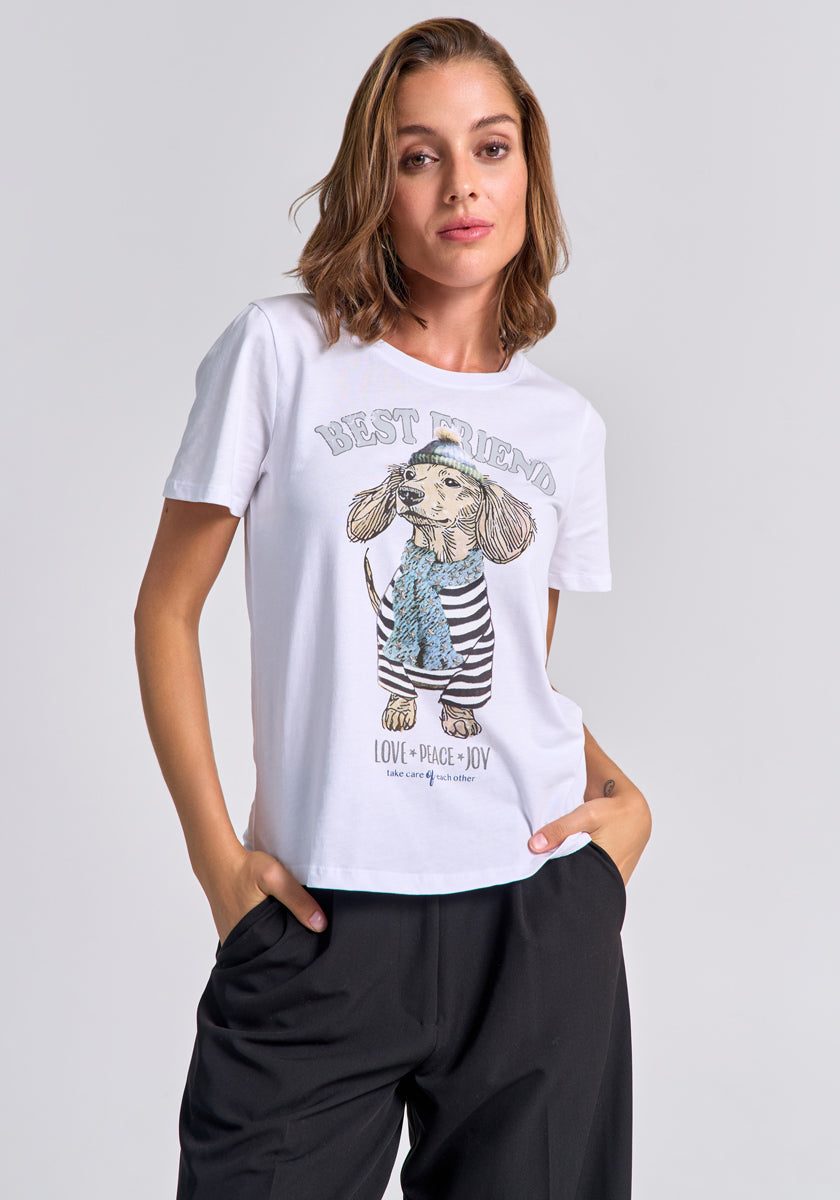 A woman with short, wavy brown hair wears a white Princess Goes Hollywood cotton T-shirt featuring a cartoon dachshund in a scarf and striped shirt. The print says “Best Friend” and “Love Peace Joy” as she stands with one hand in her black pants pocket.
