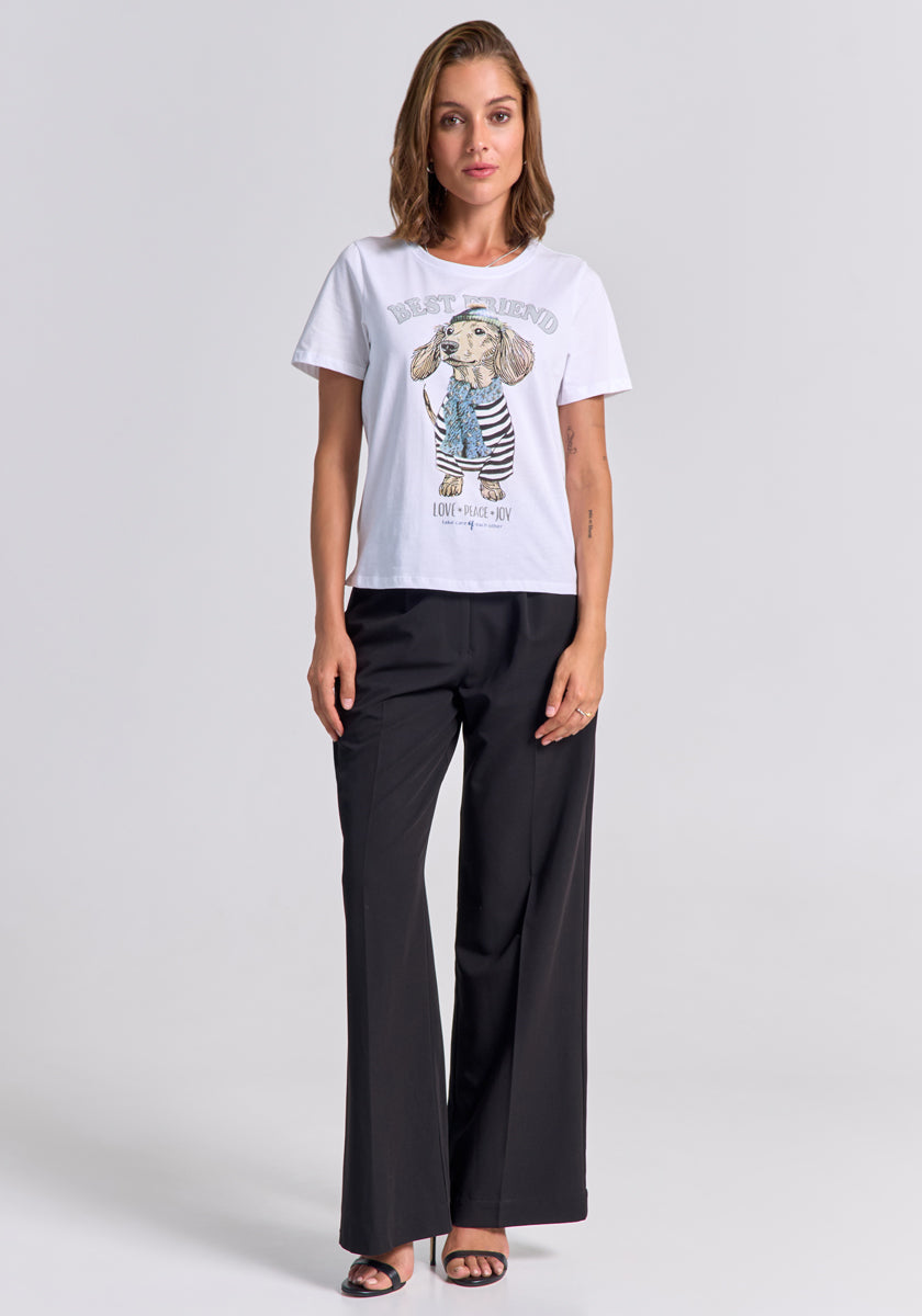 A woman with shoulder-length brown hair models a Princess Goes Hollywood white cotton T-shirt featuring a playful dachshund print in stripes and "Best Friend" text, paired with black wide-leg trousers and open-toed heels, standing before a plain light background.