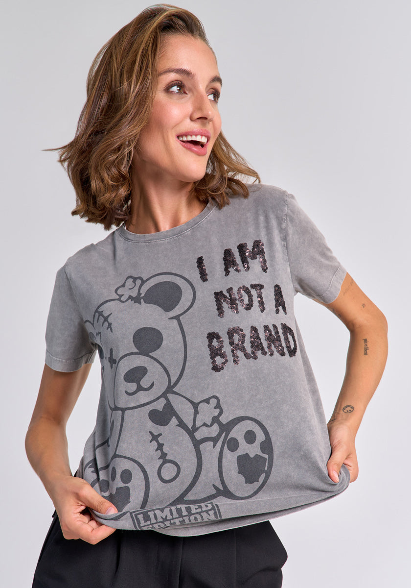 A smiling woman with shoulder-length brown hair holds out the hem of her graues Baumwoll T-Shirt from Princess Goes Hollywood, featuring a large patched teddy bear graphic and bold “I AM NOT A BRAND” text, against a plain light gray background.