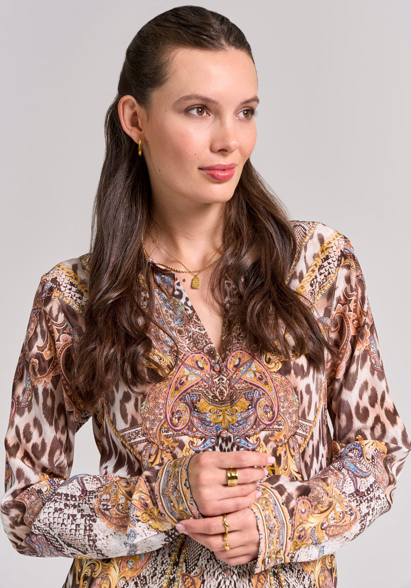 A woman with long brown wavy hair wears a Princess Goes Hollywood Snake-Leo-Print Seidenbluse, gold hoop earrings, rings, and a necklace. She stands against a neutral background, gazing slightly right with a gentle expression.