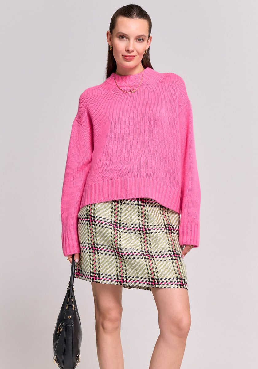 A woman stands against a plain background, wearing a bright pink knit sweater and a Karo-Print flared skirt with red and black accents from Princess Goes Hollywood. She carries a black handbag, her brown hair tucked behind her ears as she smiles softly.