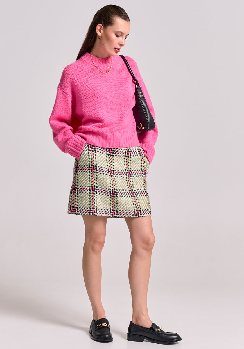 A woman with long brown hair stands against a plain background wearing a bright pink Princess Goes Hollywood sweater, an A-line check skirt with pockets, black loafers, gold jewelry, and a black shoulder bag. Her hands rest in her skirt pockets.