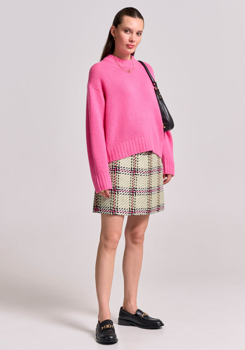 A woman stands against a plain light background wearing a loose pink sweater, a Karo-Print Minirock aus Tweed mit Gürtelschlaufen from Princess Goes Hollywood, black loafers with gold hardware, and a black shoulder bag. Her sleek hair is tucked back.