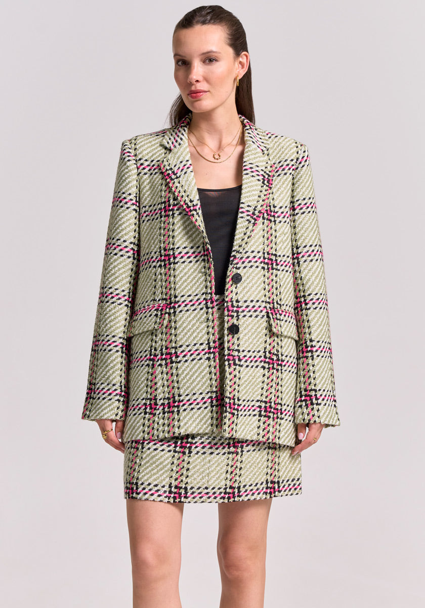 A woman with pulled-back brown hair confidently faces the camera in a Princess Goes Hollywood Karo-Print Blazer with pink and black accents over a black top, paired with a Tweed Minirock featuring Gürtelschlaufen, against a light background.