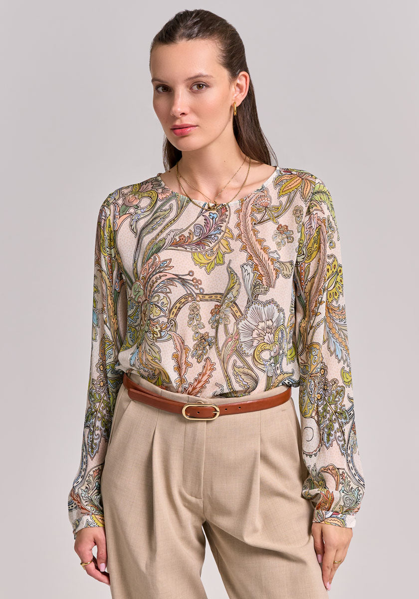 A woman with straight brown hair wears a beige paisley-print blouse with long sleeves by Princess Goes Hollywood, tucked into high-waisted tan pleated trousers with a slim brown belt, standing against a gray background.