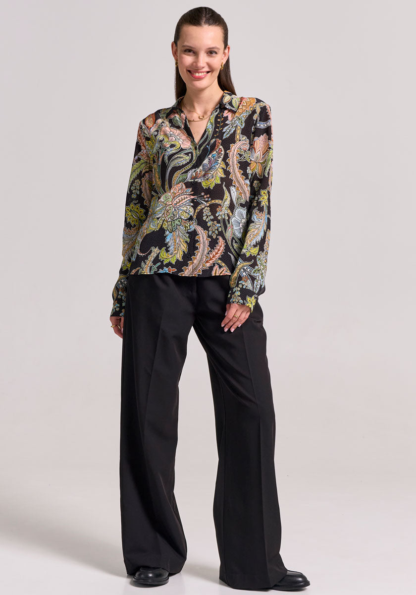A woman smiles, wearing a long-sleeved black Paisley-print Hemdbluse and black wide-leg trousers from Princess Goes Hollywood. She pairs them with shoes and a necklace; her hair is slicked back. The background is light gray.