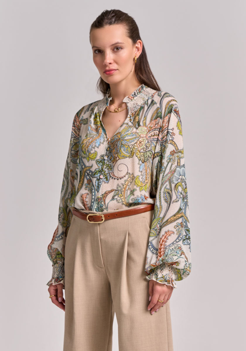A woman with long brown hair pulled back wears a Paisley-Print Bluse with Balloon sleeves and a smocked stand-up collar from Princess Goes Hollywood, tucked into high-waisted beige trousers, accessorized with gold jewelry and a brown belt.