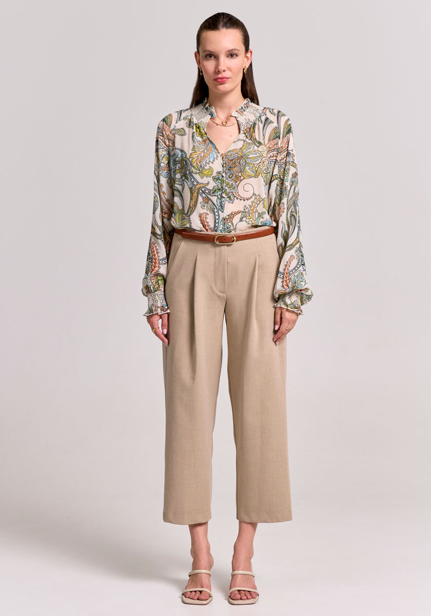 A woman faces forward against a light gray background, wearing a Princess Goes Hollywood paisley-print blouse with balloon sleeves and smocked stand-up collar, tucked into high-waisted beige ankle-length pants with a brown belt.